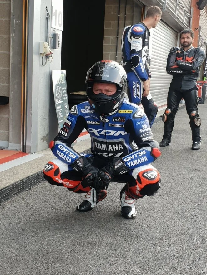 Un motard en tenue de course avec un casque, plie le genou sur le côté, en dehors d'une écurie de course. Deux autres personnes en tenue de course se tiennent à l'arrière, face à lui, devant un local à volet roulant métallique.