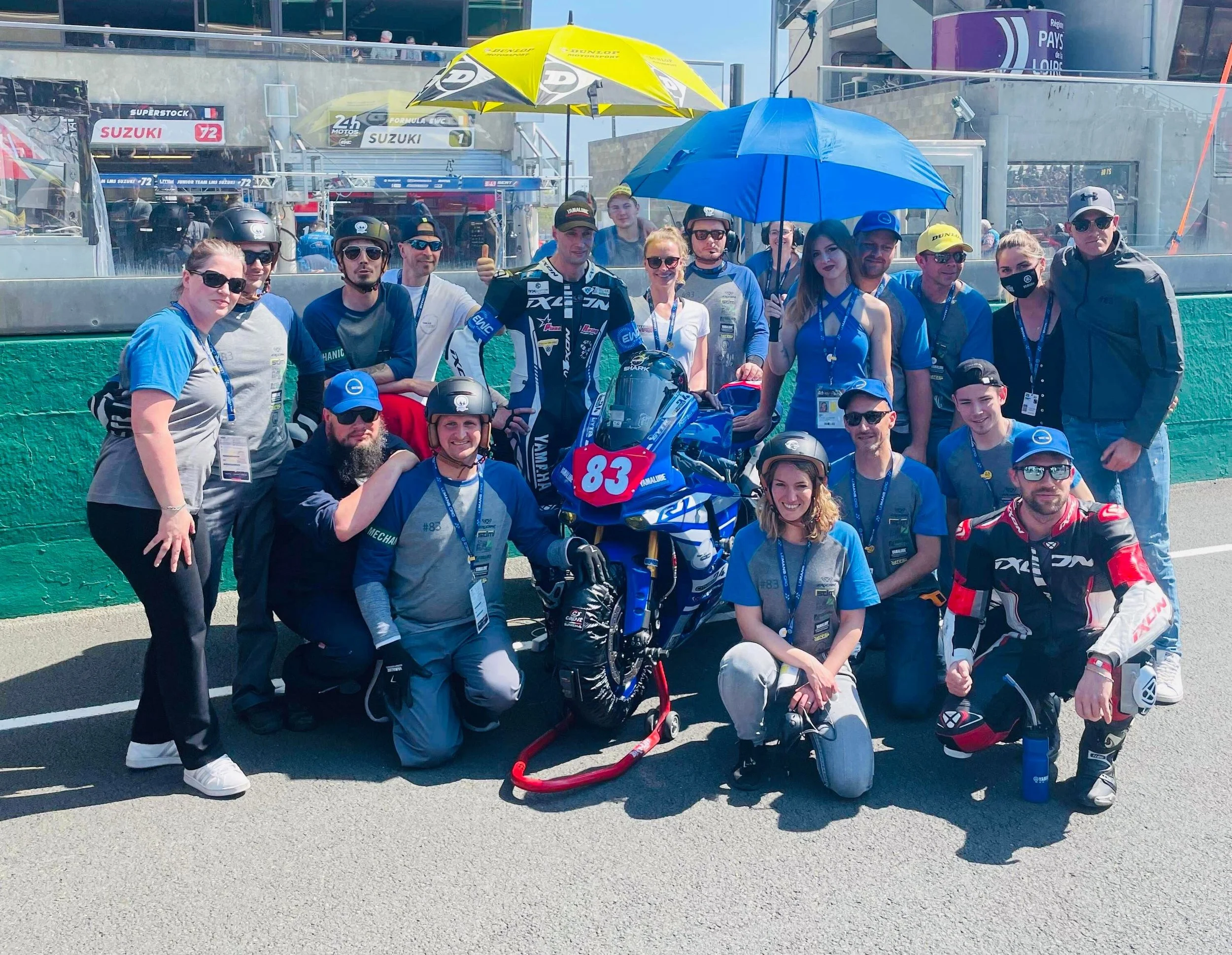 Groupe de gens autour d'une moto de course numéro 83 sur un circuit de course. Certains portent des casques et des vêtements de course, d'autres portent des t-shirts avec des badges. Il y a des parapluies pour se protéger du soleil.