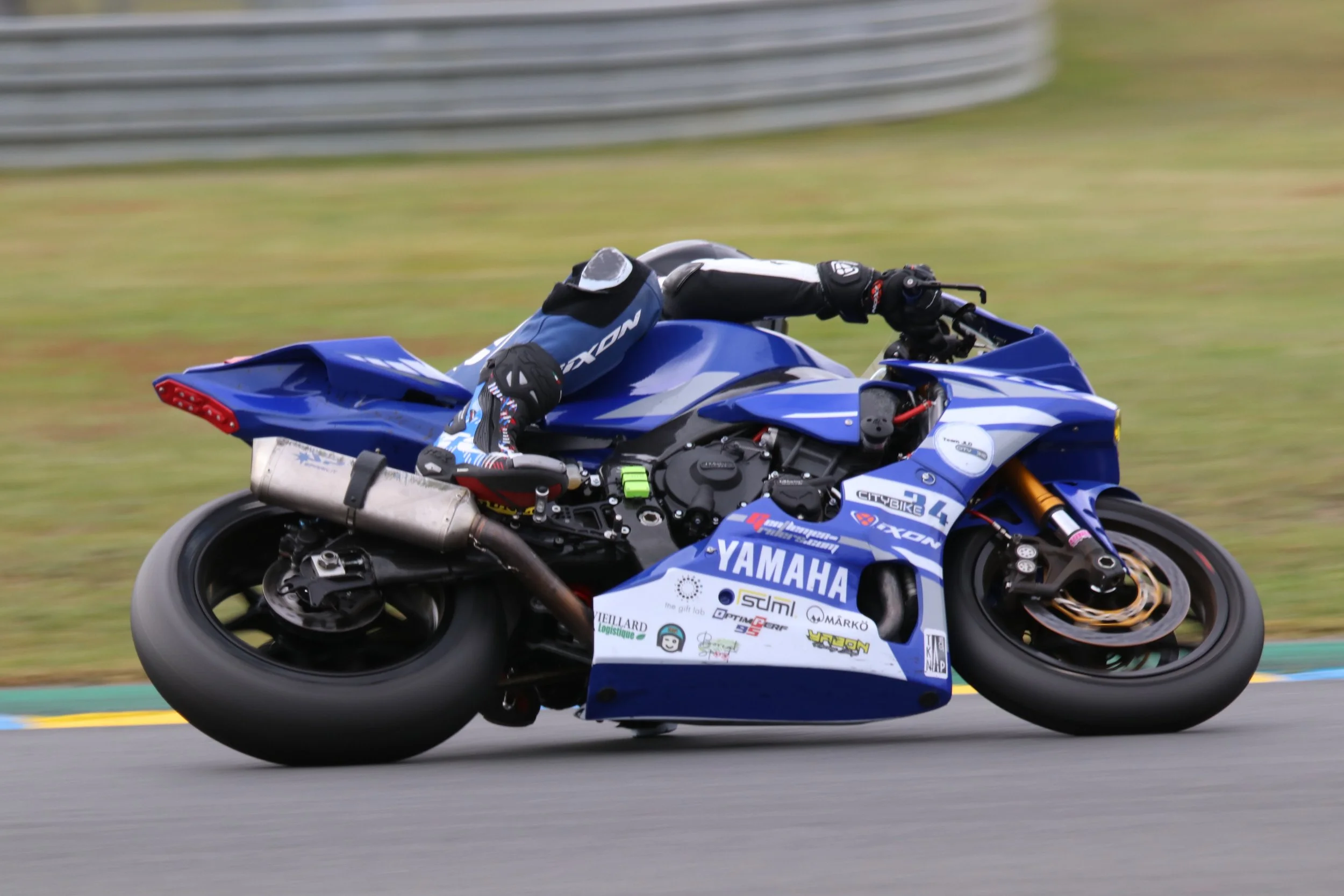 Une moto de course Yamaha bleue en plein virage sur une piste de course, avec un pilote en combinaison de course noire et bleue et casque noir, floue derrière pour indiquer la vitesse.