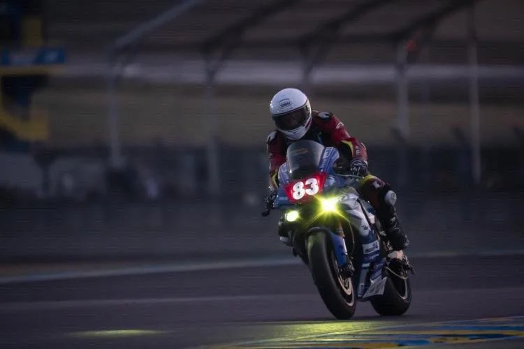 Un pilote de moto en course, portant un casque blanc et un équipement rouge, roulent à grande vitesse sur une piste de course, avec le numéro 83 sur la moto et les phares allumés.