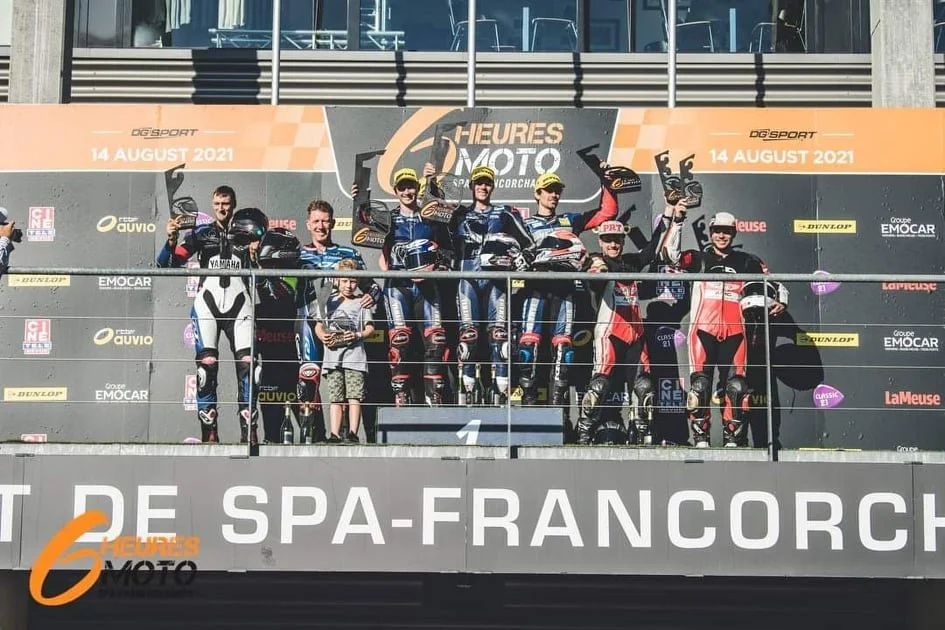 Groupe de pilotes de moto sur le podium de la course, célébrant leur victoire lors d'une compétition de moto à Spa-Francorchamps, avec palmarès et sponsors affichés en arrière-plan.