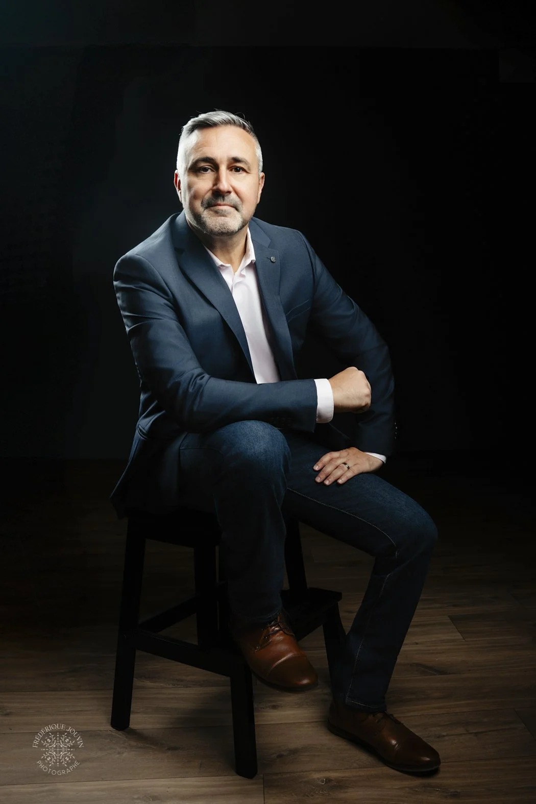 Un homme d'âge moyen en costume bleu et pantalon foncé, assis sur une chaise en bois, pose devant un fond noir. Il a des cheveux grisonnants, une barbe courte, et porte une chemise blanche. Il look sérieux et professionnel.