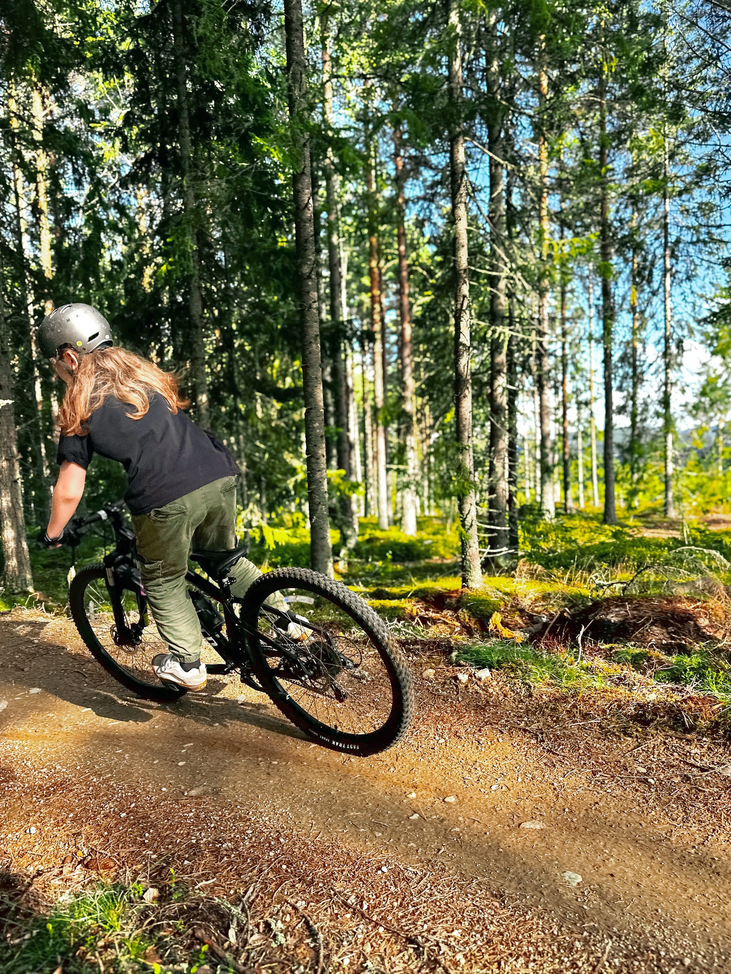 Person som cyklar på en stig genom en skog med grön träd och blå himmel i bakgrunden.