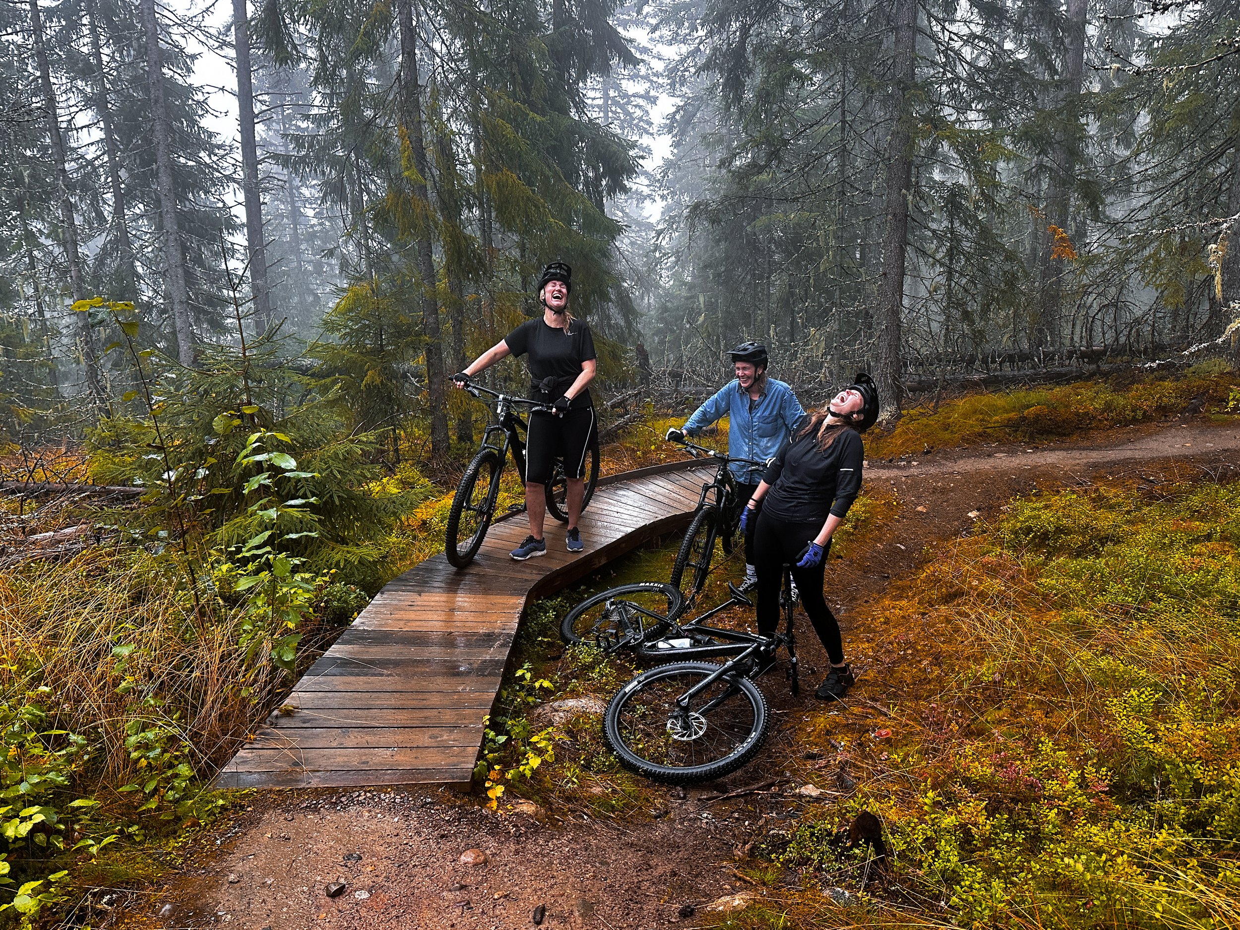 Tre personer med cyklar som skrattar och ler på en liten träbro i en dimmig skog.