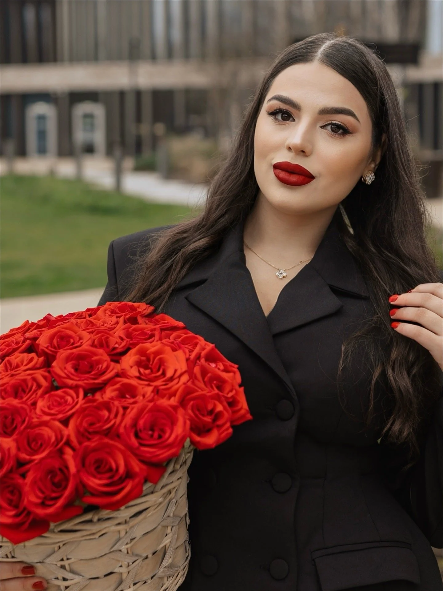 A woman with long dark hair, red lipstick, and a black blazer holding a basket of red roses outdoors.