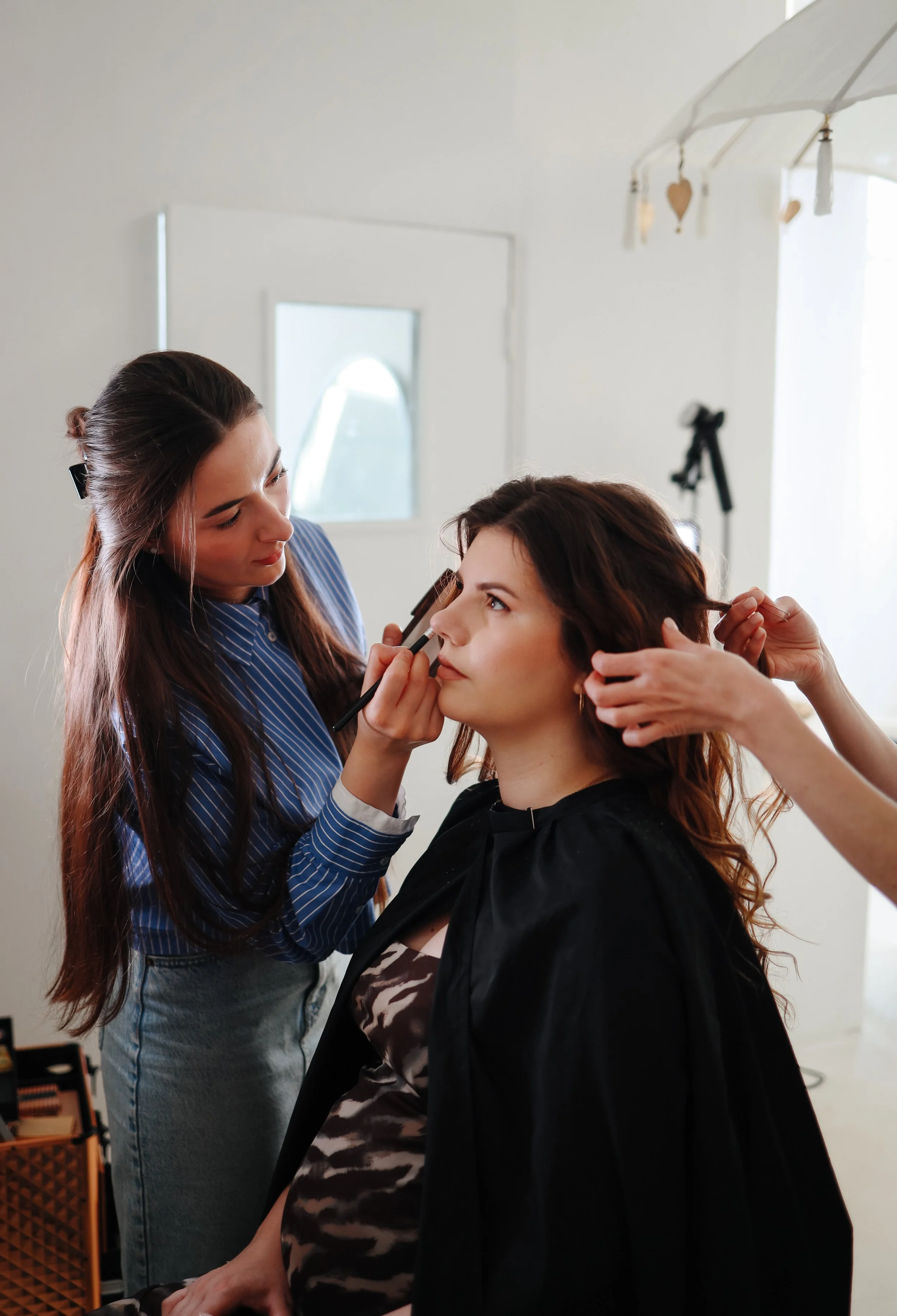 A makeup artist applying eye makeup to a woman sitting in a chair, with another person helping with her hair in a well-lit indoor space.