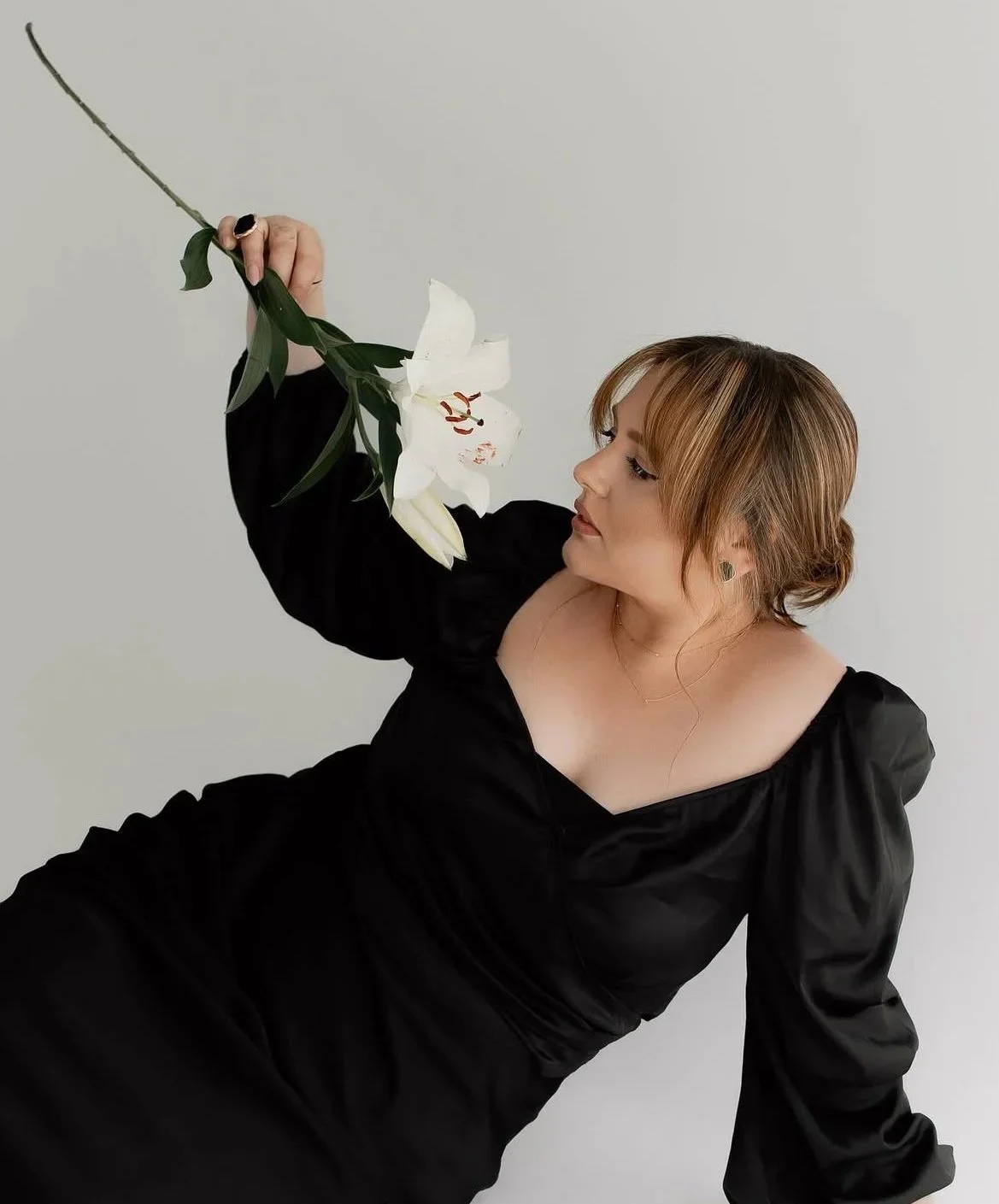 A woman in a black dress holding a white lily flower in the air while looking at it.