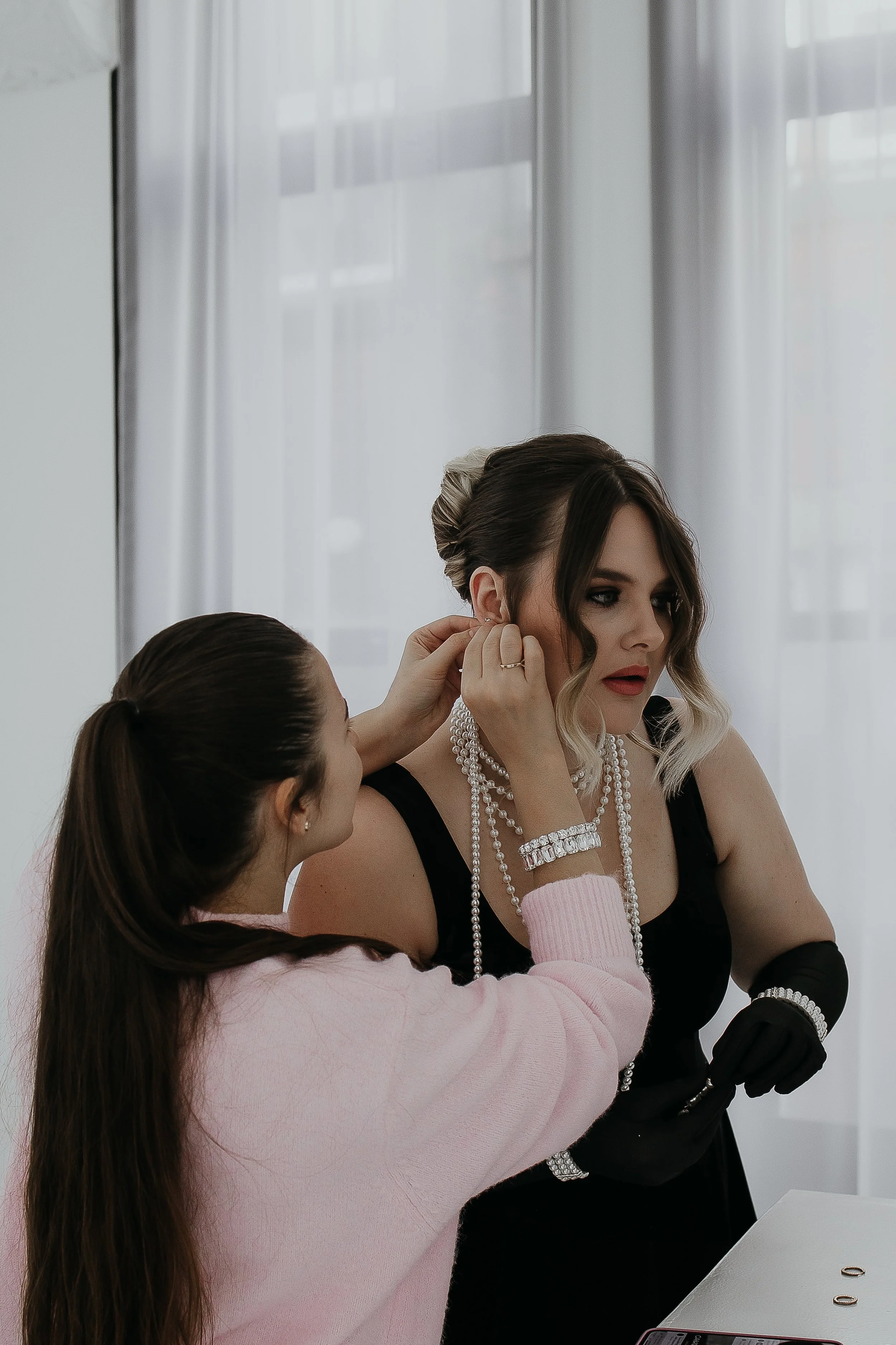 A woman with styled hair and jewelry is being assisted with her earrings by another woman in a pink sweater, in a well-lit room with white curtains.