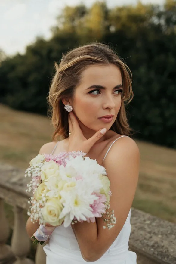 A young woman with light brown hair in loose waves wearing a white dress with thin straps, holding a bouquet of white and pink flowers, standing outdoors with trees and a stone railing in the background.