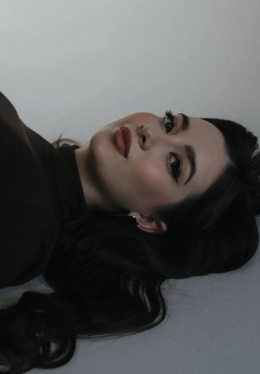A woman with long dark hair and makeup, wearing a black top, looking at the camera with a neutral expression against a plain light-colored wall.