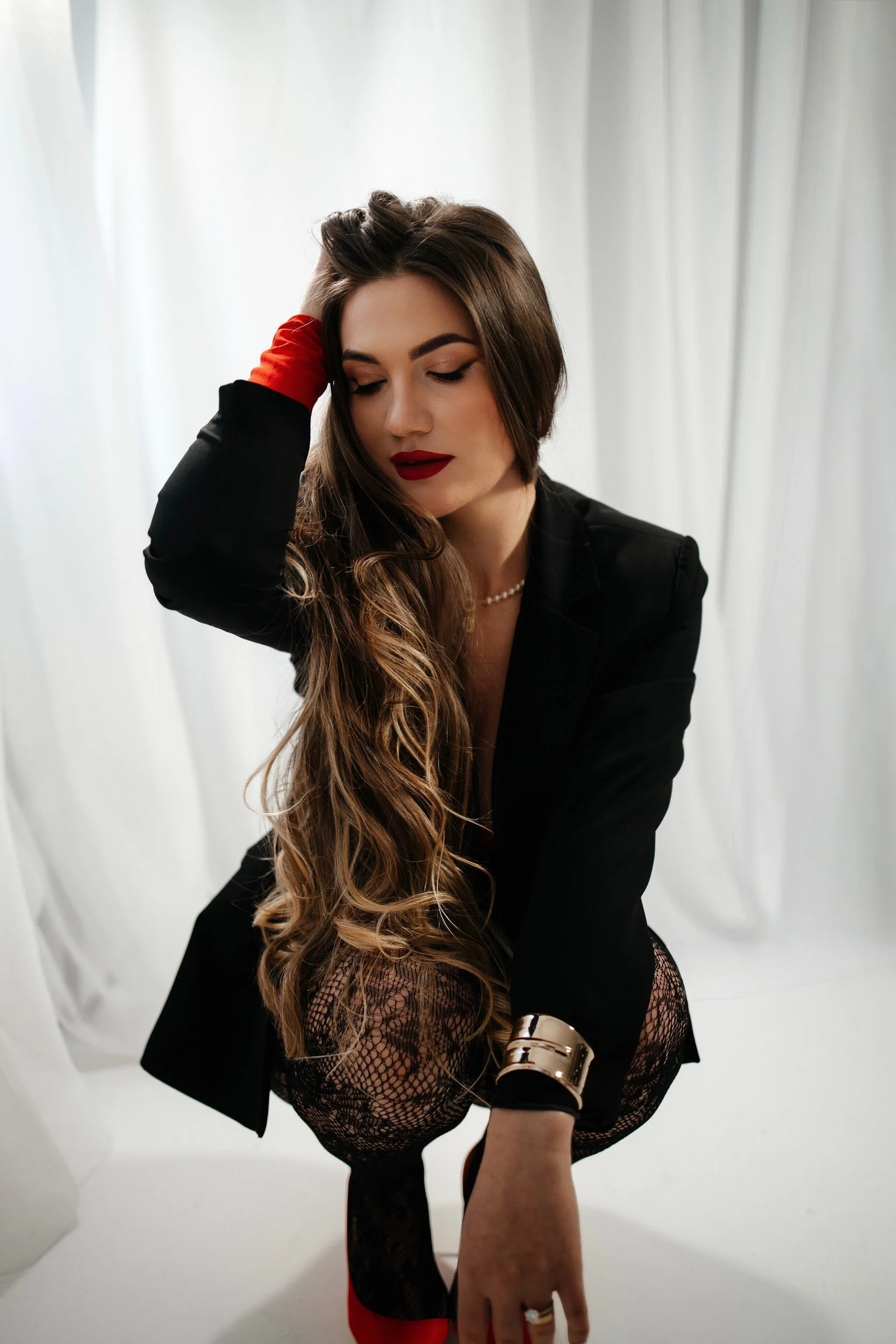 A woman with long wavy hair, wearing a black blazer, red lipstick, and black lace stockings with red heels, is posing with her eyes closed, touching her hair and ankle, against a white background.