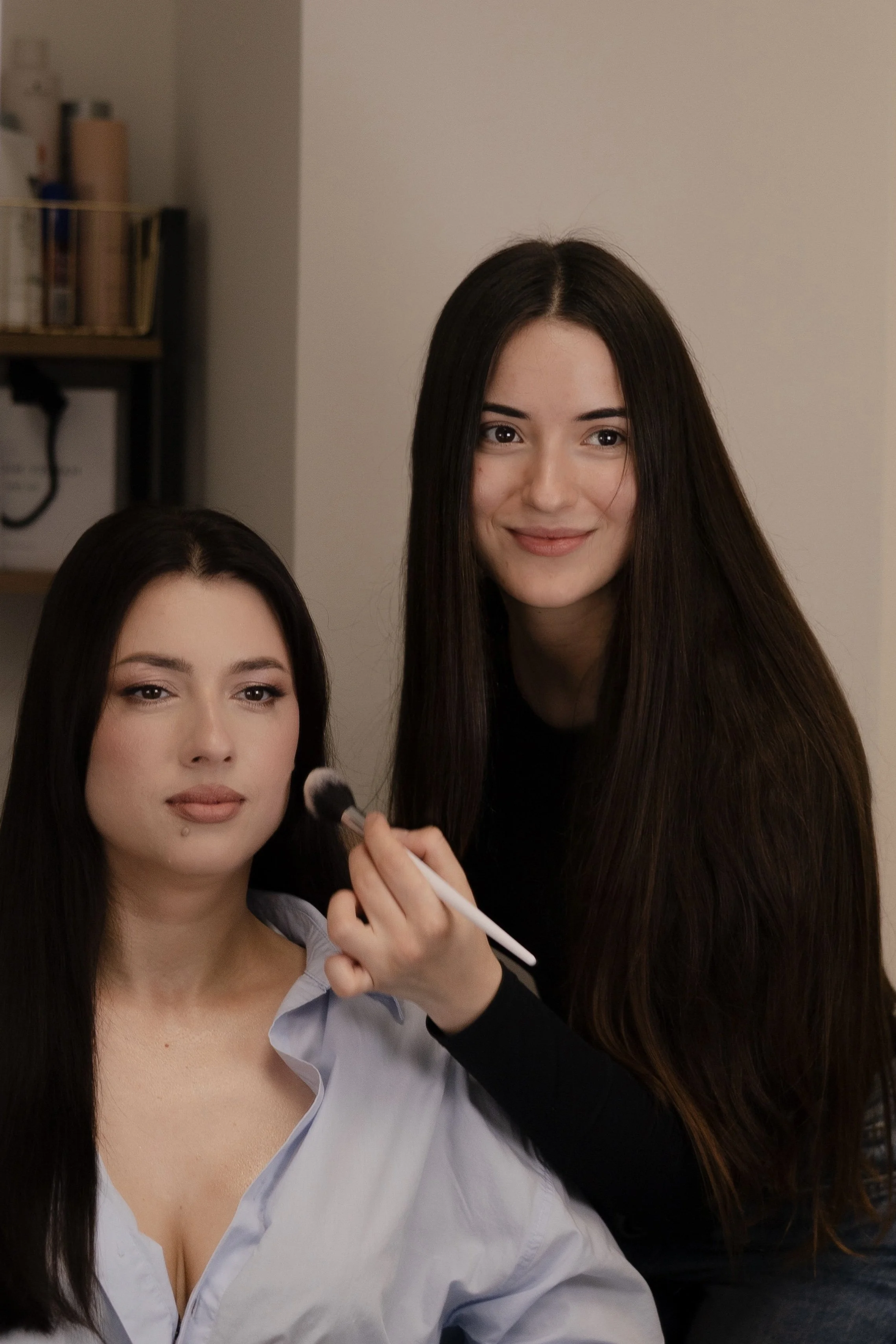 Two women, one sitting and one standing, with the woman sitting applying makeup while the other assists.