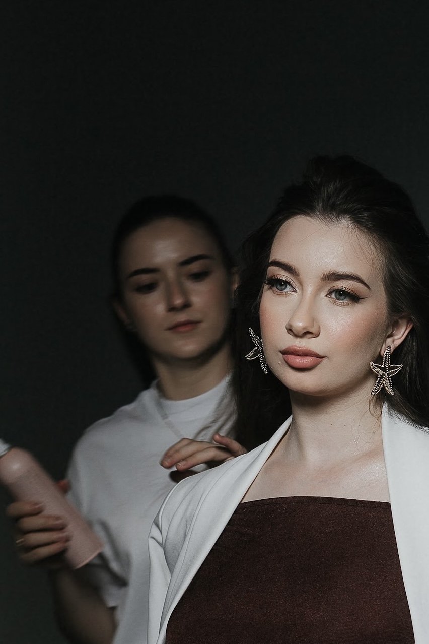 Stylish woman with starfish-shaped earrings, wearing a dark top and white blazer, poses in front of a dark background while another woman applies makeup or adjusts her hair behind her.