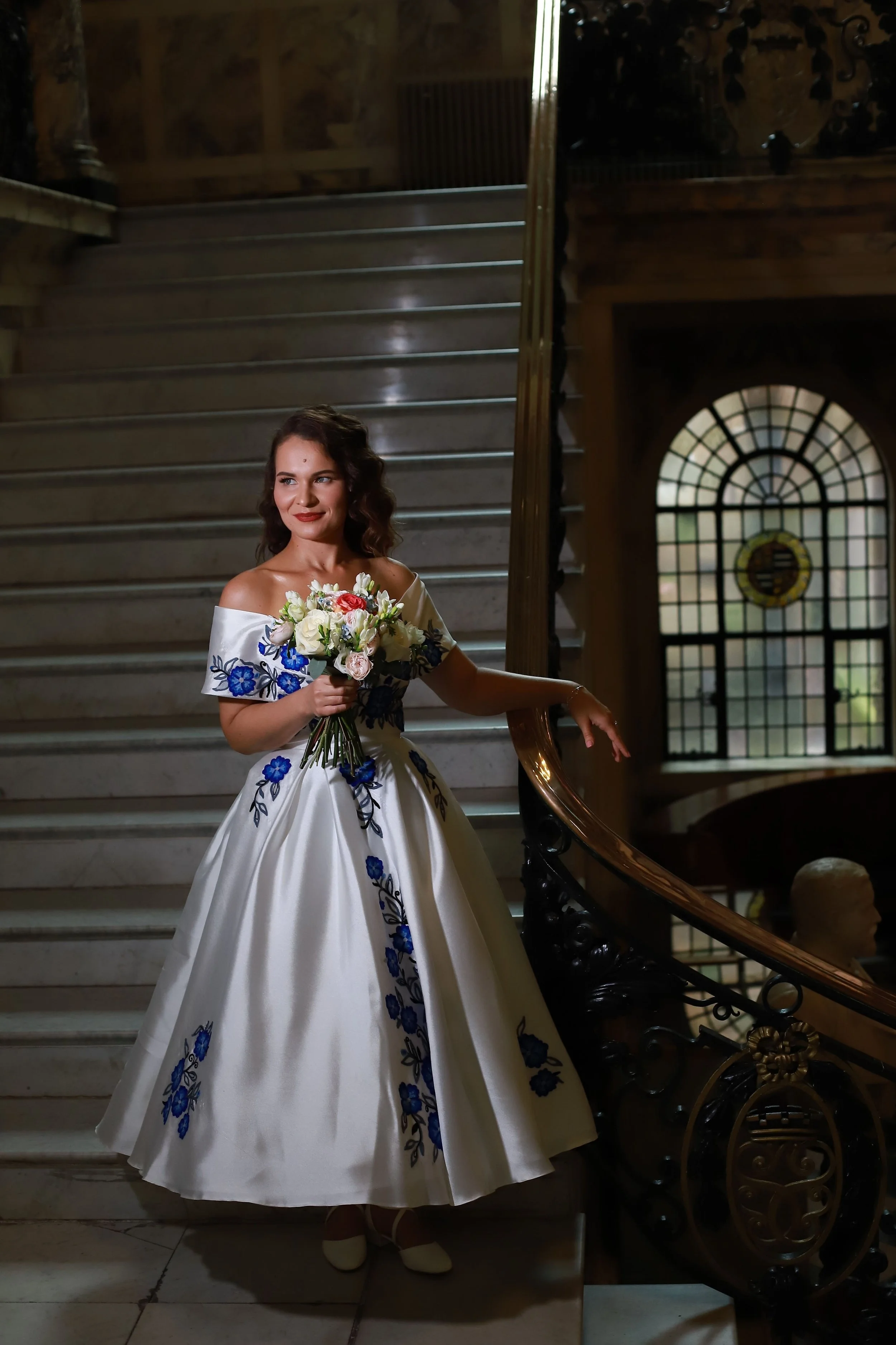 A woman in a white wedding dress with blue floral embroidery holding a bouquet of white and pink flowers, standing on a staircase in a historic or elegant interior, with a stained glass window in the background.