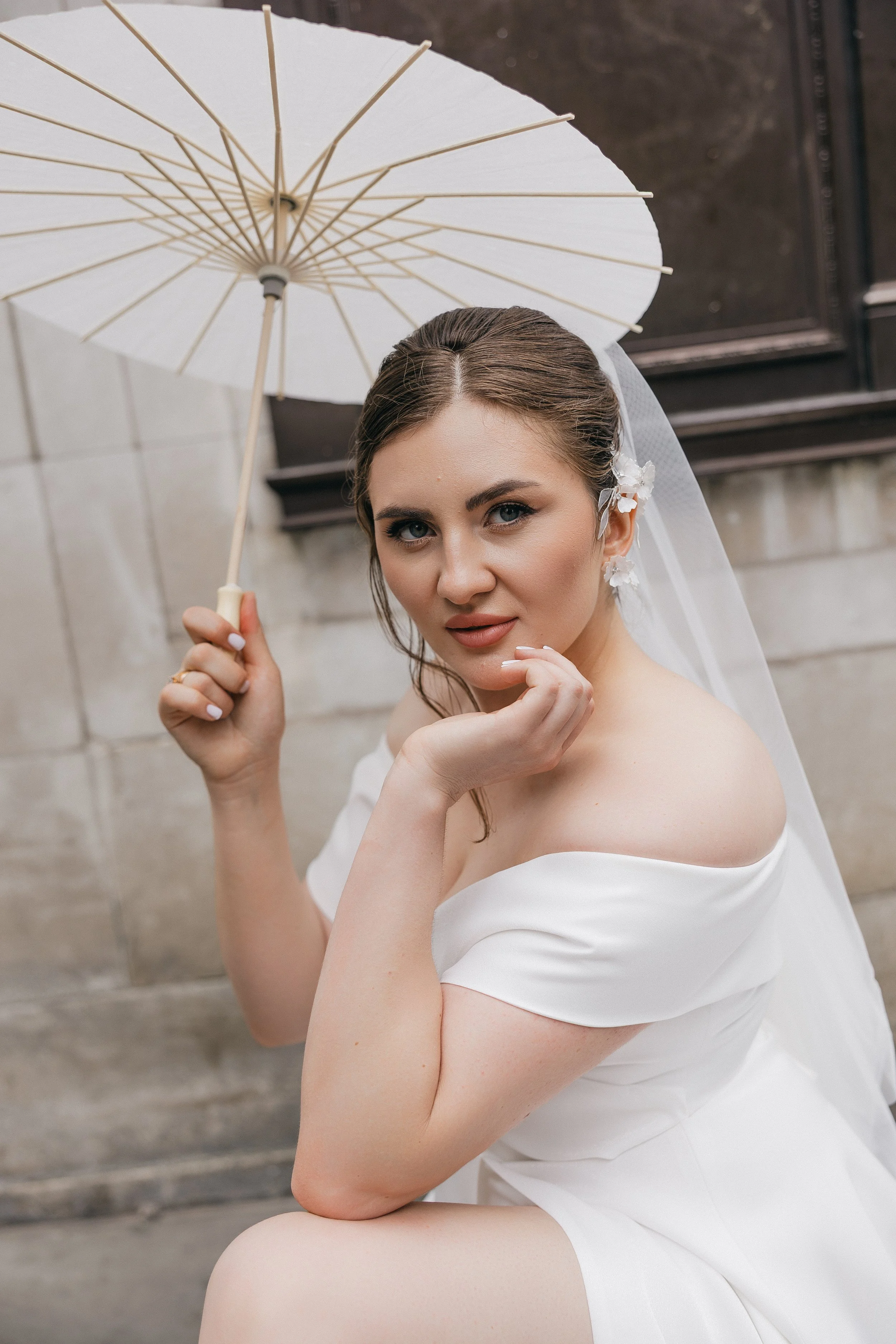 Portrait of a bride holding a white umbrella, wearing an off-the-shoulder white wedding dress and a veil, standing outdoors near a stone wall.