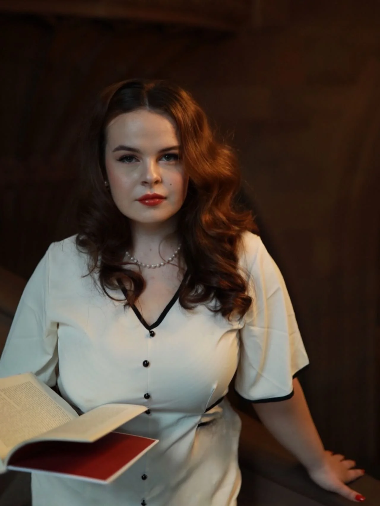 A woman with wavy brown hair, dressed in a cream blouse with black trim, sitting with an open book. The background is dark and the lighting highlights her face and upper body.