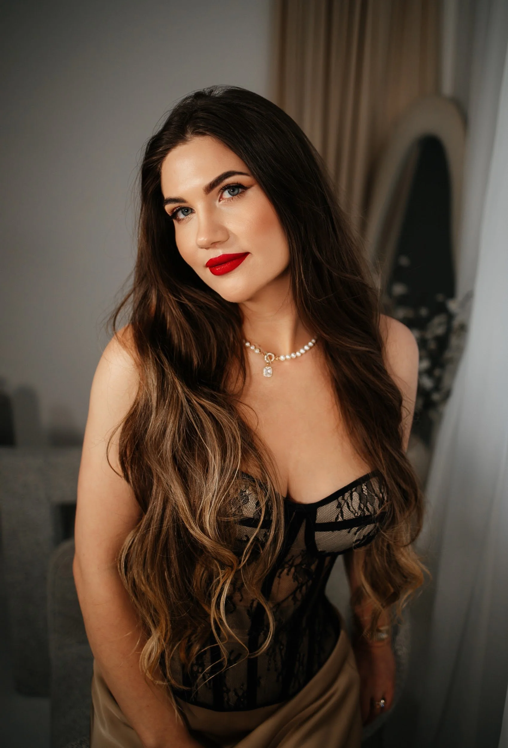 A woman with long wavy brown hair, red lipstick, and blue eyes, wearing a black lace corset-style top and a pearl necklace, stands indoors near a mirror and curtain.