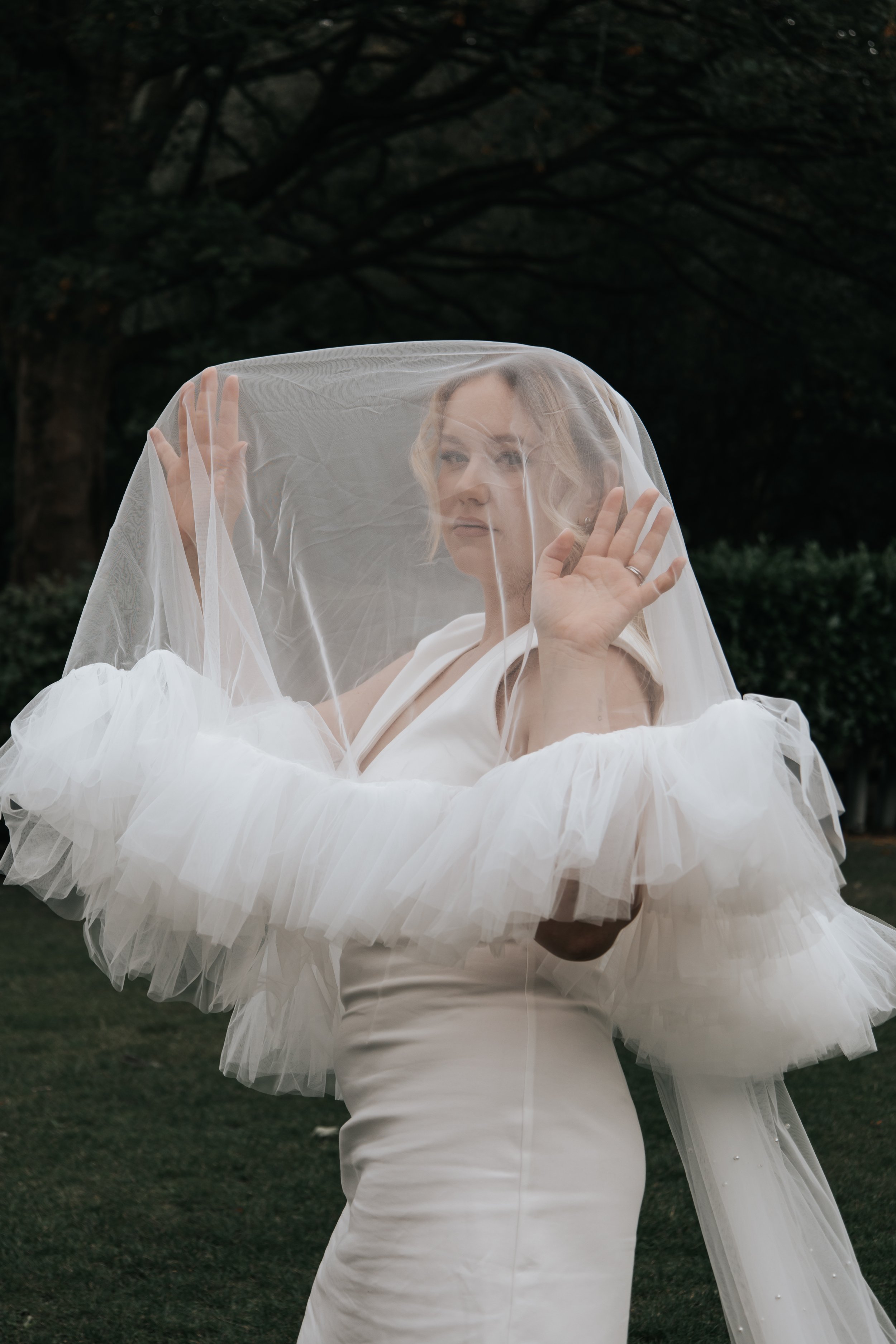 A woman in a white wedding dress with ruffled sleeves holding a sheer veil, standing outdoors with dark trees in the background.