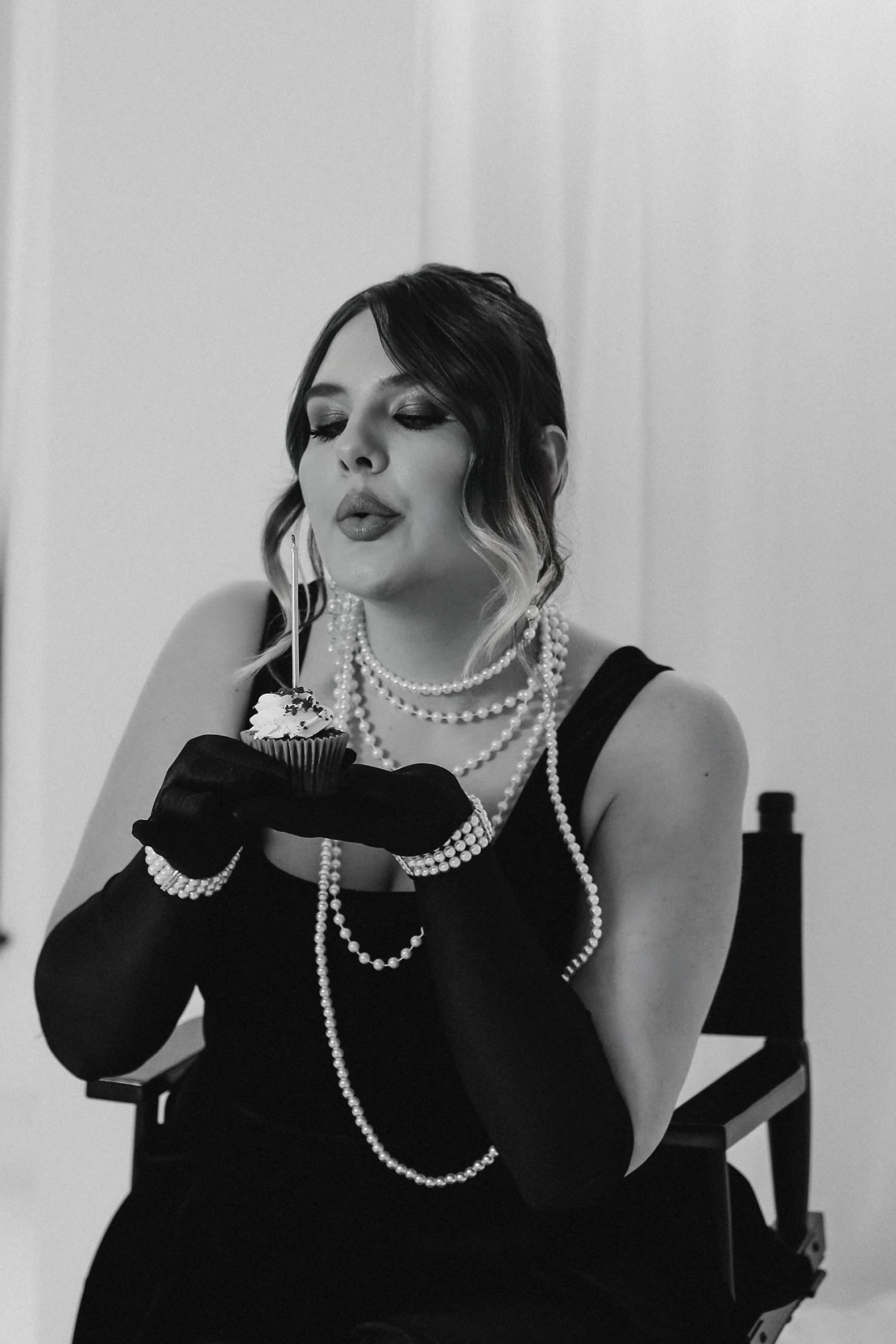 A woman with dark hair and makeup, dressed in black with pearl jewelry and long black gloves, is blowing out a candle on a cupcake while sitting on a chair.