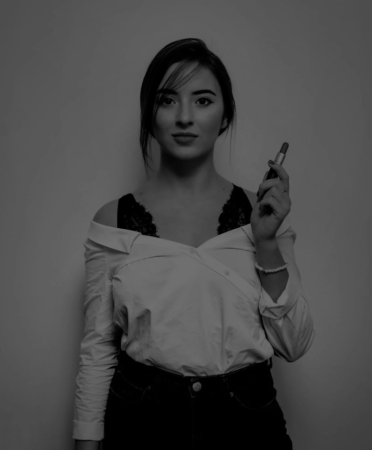 Black and white photo of a woman holding a lipstick, wearing an off-shoulder shirt with lace detail underneath and dark pants.