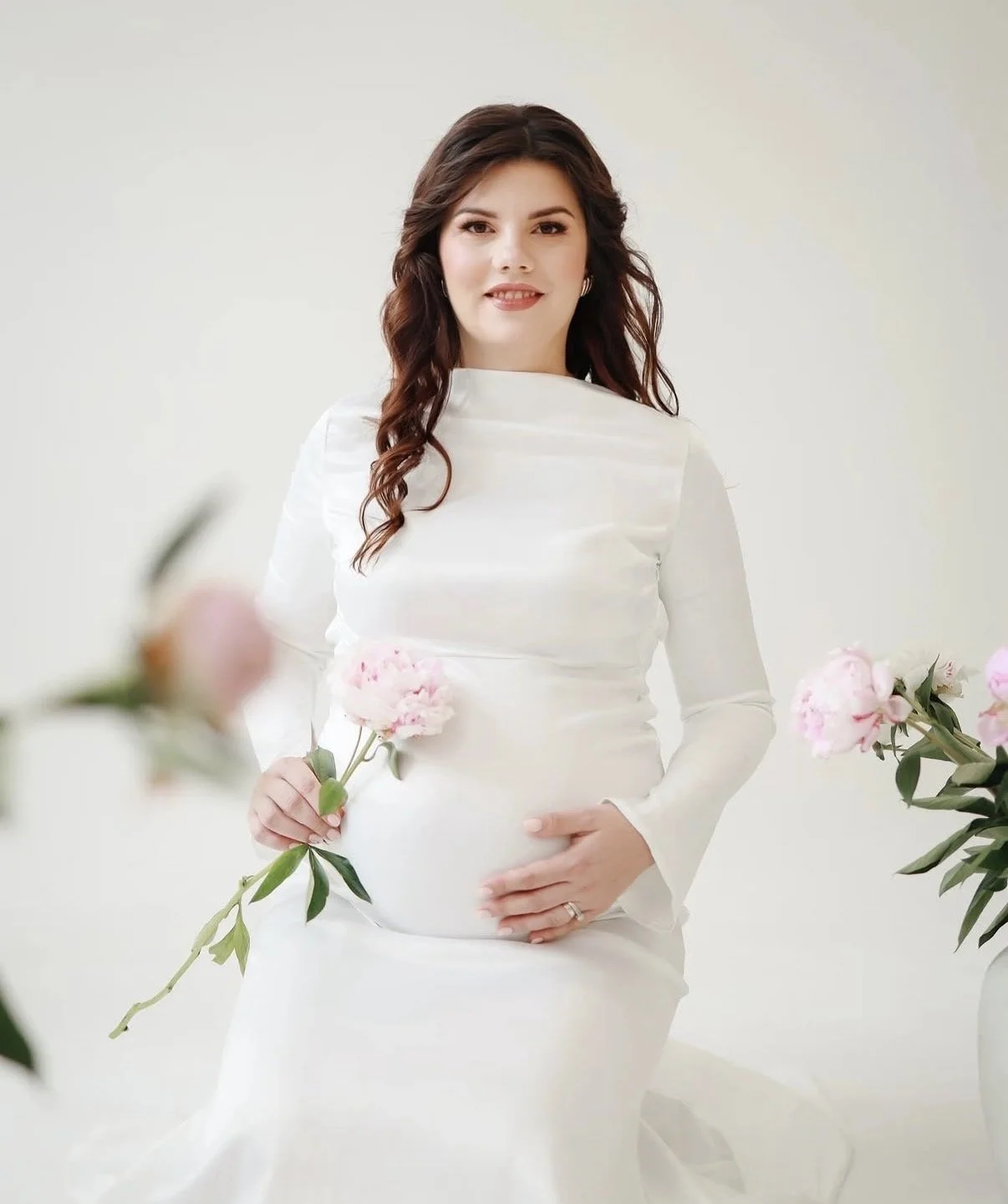 A pregnant woman in a white dress holding pink flowers, standing in front of a plain light-colored background.