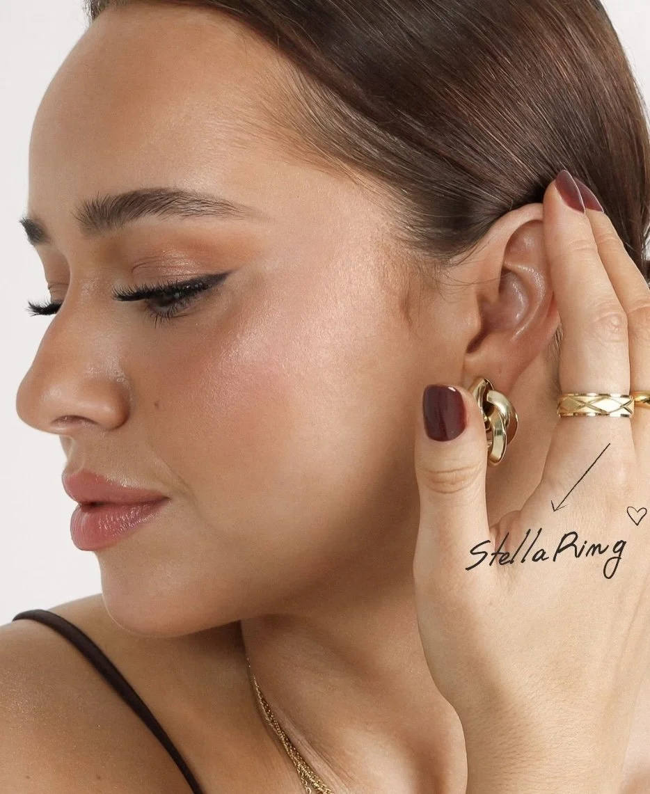 A woman with brown hair, wearing gold jewelry including a hoop earring and rings, is touching her ear. The photo highlights a gold ring labeled 'Stella Ring' on her finger.