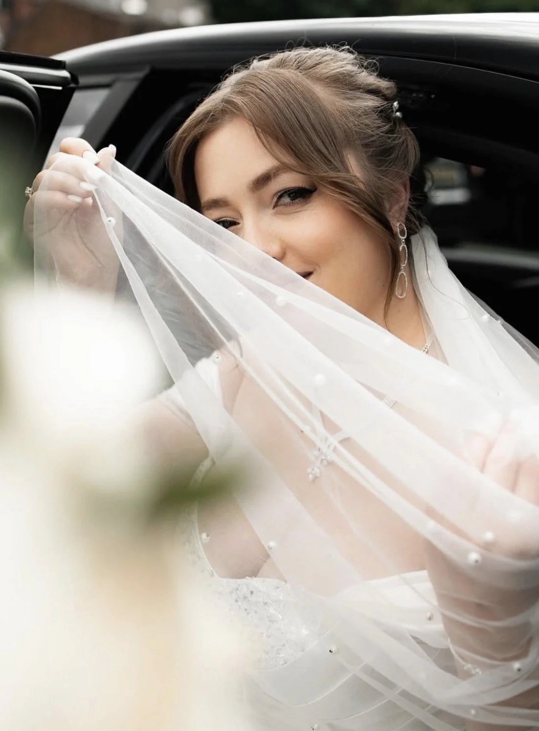 Bride in a wedding dress partially covering her face with her veil, smiling, with jewelry, standing near a black vehicle.