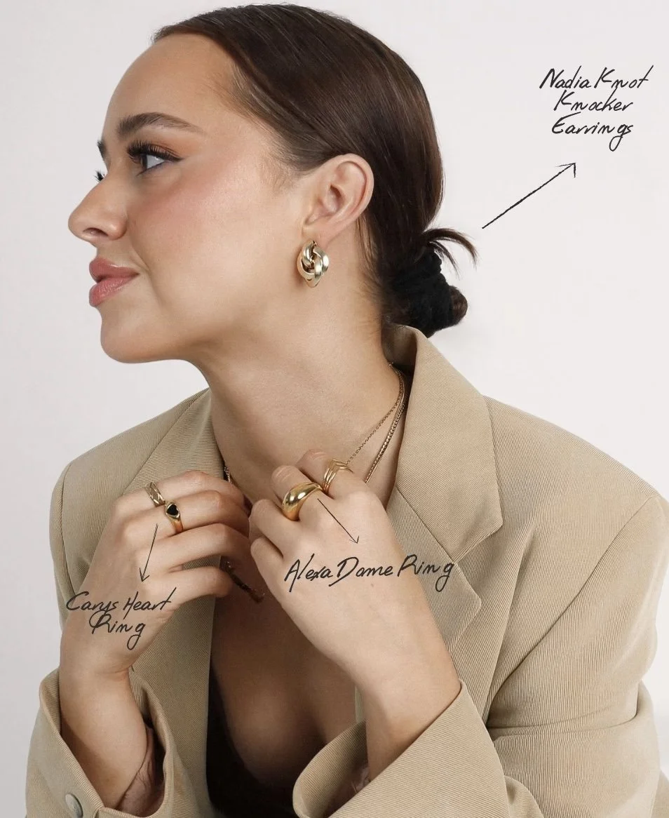 A woman with dark brown hair tied back, wearing a beige blazer, multiple gold jewelry pieces including Nadia Knot Knocker Earrings, Alexa Dame Ring, and Carpe Heart Ring, smiling and looking to the left.