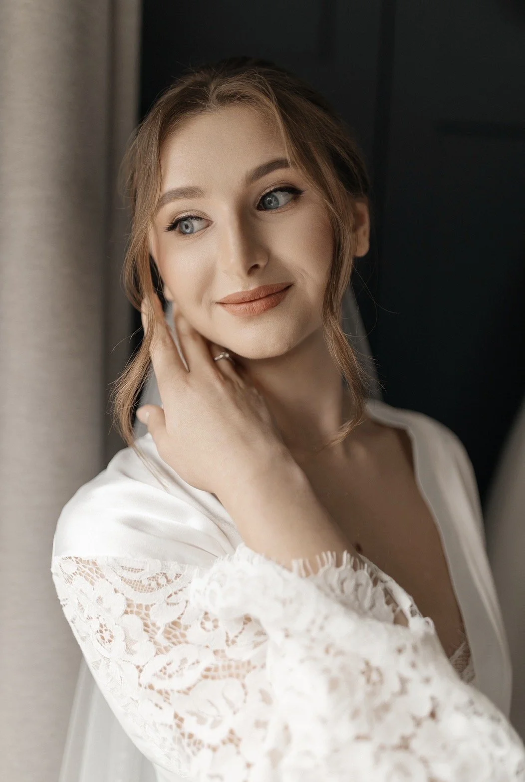 A woman with red hair and blue eyes looking to the side and touching her face with her hand, wearing a white lace top.