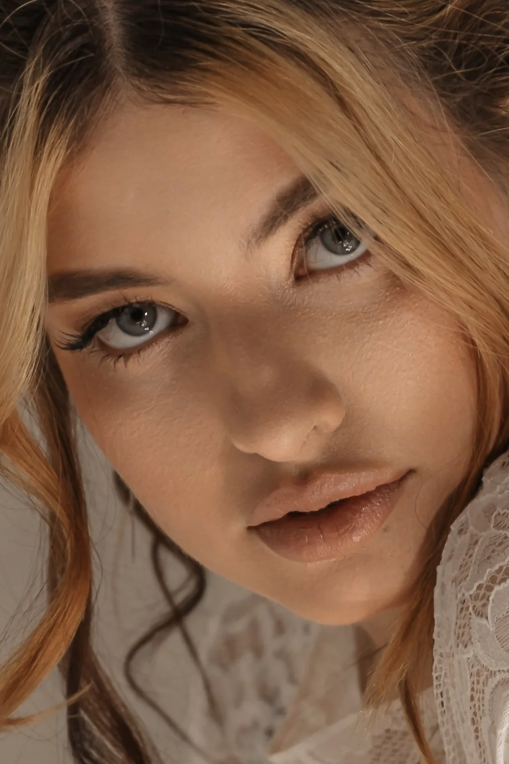 Close-up of a woman's face with light skin, blonde hair, gray eyes, and wearing makeup, in a lace garment.