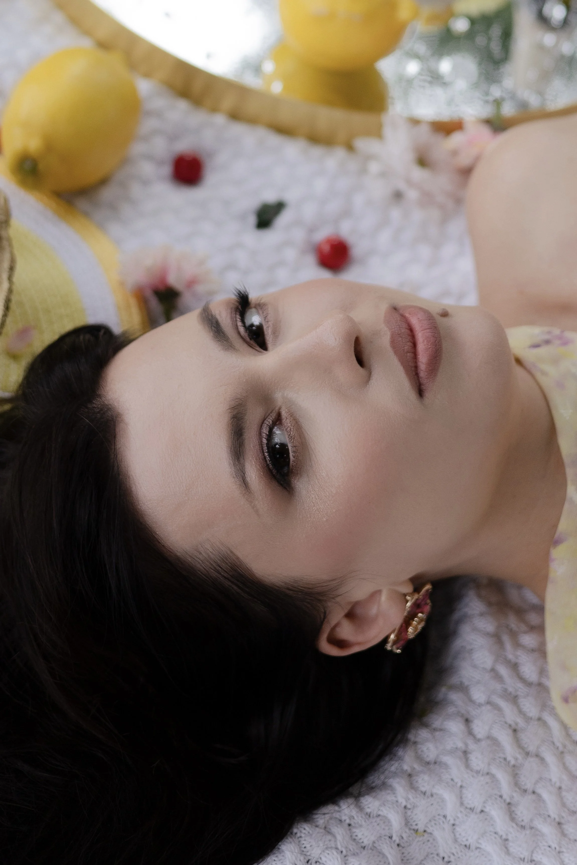 A woman with dark hair and makeup lying on a textured white surface, surrounded by yellow lemons, pink flowers, and small red berries.