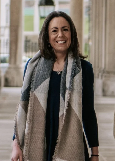 A woman with shoulder-length dark brown hair, wearing a dark blue top, a gray scarf, and a black bracelet, smiling in front of a background with classical architectural columns and large windows.