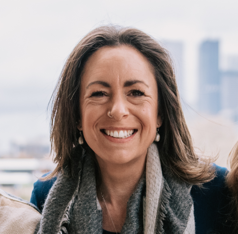 A woman smiling outdoors with a city skyline in the background.