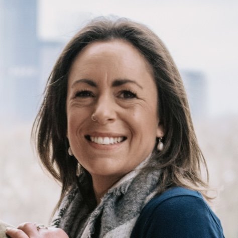 Smiling woman with shoulder-length brown hair, wearing a gray scarf, earrings, and a blue top, indoors with a window in the background.