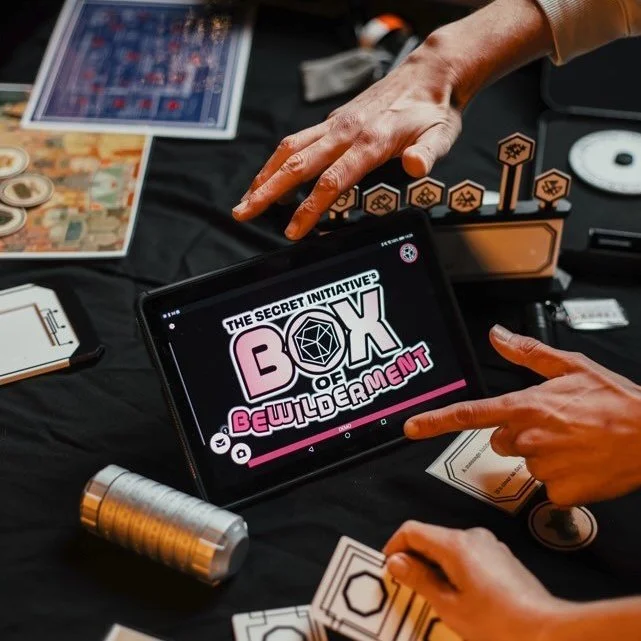 Two people playing a tabletop game called 'Box of Bewilderment' from The Secret Initiative, with game components, tokens, cards, and a tablet displaying the game title, on a black table.