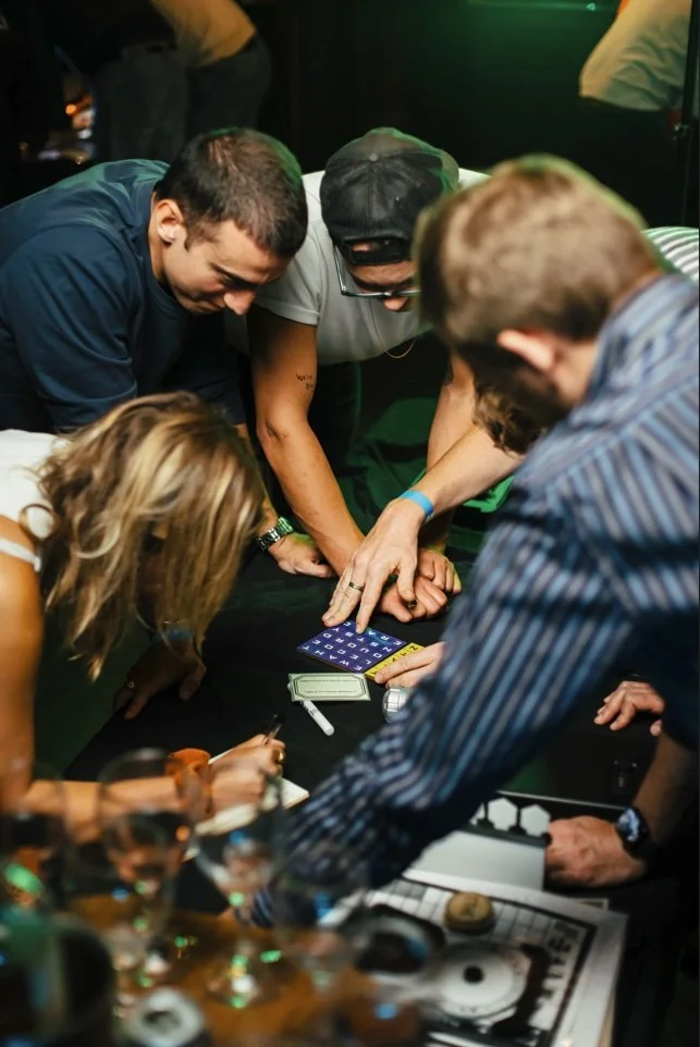 Group of people playing a game together at a table, with cards, money, and game pieces visible.
