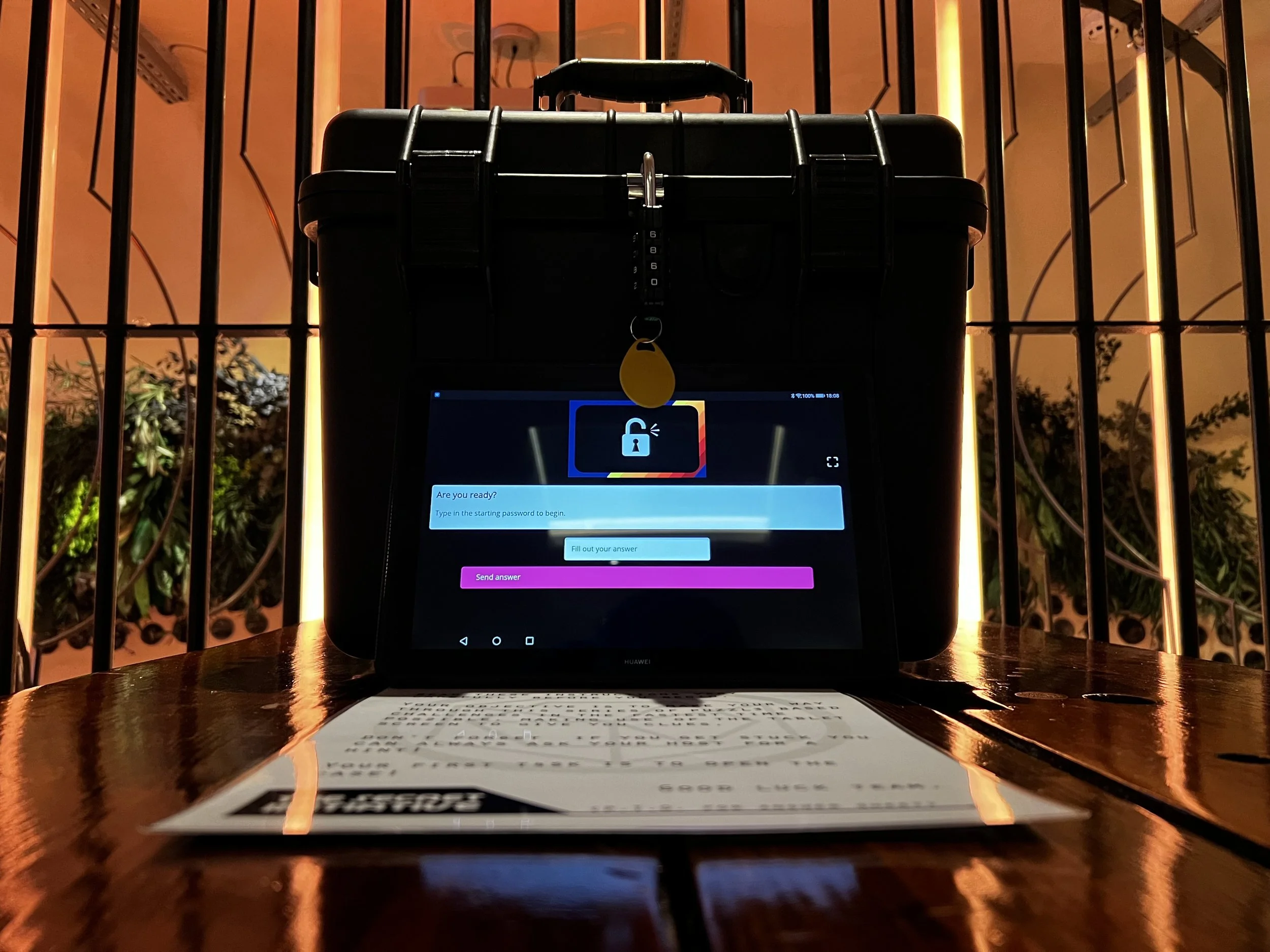 A tablet on a wooden table displays a lock screen with a message 'Are you ready? Type in the starting password to begin.' The tablet is positioned in front of a black case or bag with a combination lock, and there are vertical black metal bars and some green plants in the background.