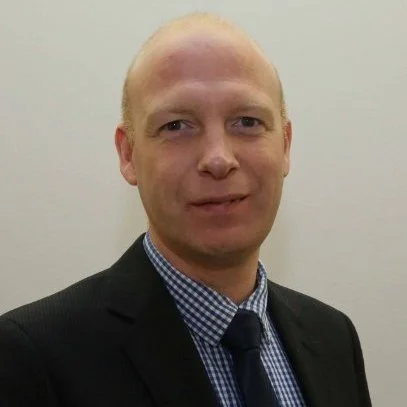 A man with light skin and a bald head wearing a black suit, checkered shirt, and black tie, standing against a plain light-colored wall.