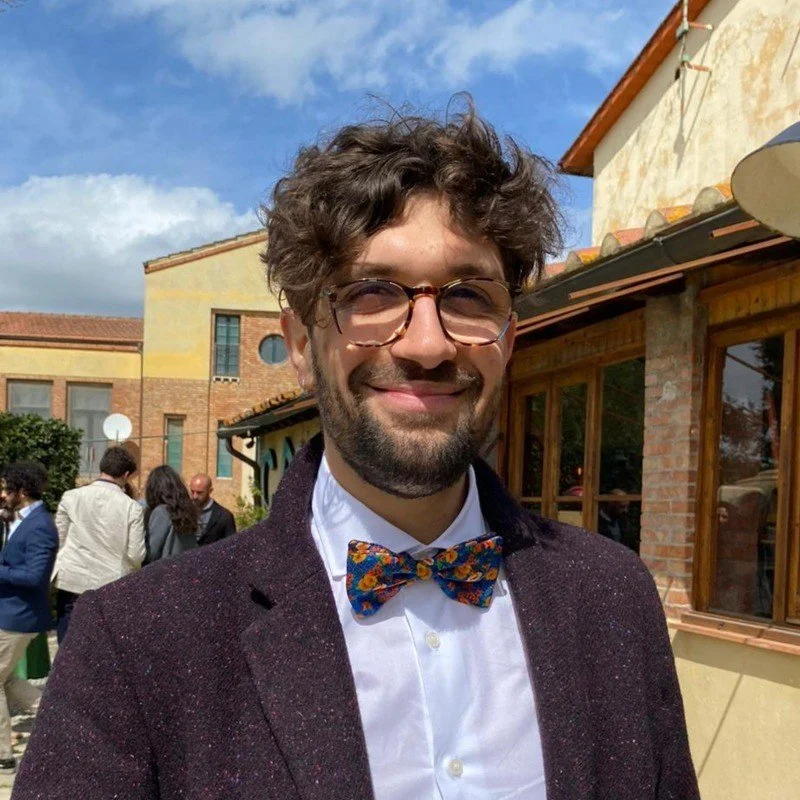 A young man with glasses and a beard smiling outdoors, wearing a white shirt and a colorful bow tie, in front of a building with other people in the background.