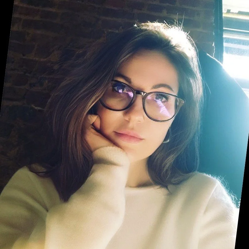 A young woman with shoulder-length brown hair and glasses, sitting indoors near a window with sunlight, resting her face on her hand, wearing a light-colored sweater.