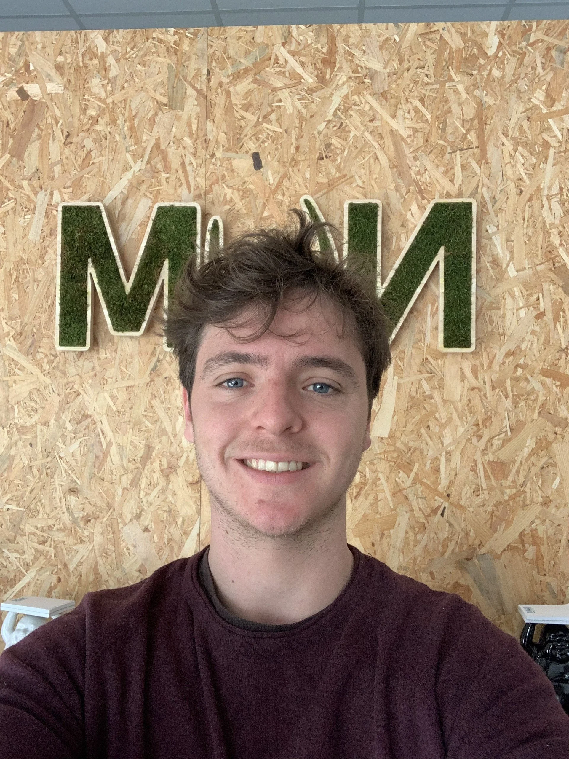 A young man with brown hair and blue eyes smiling in front of a wooden wall with the letters 'WOM' in green moss-style decoration.