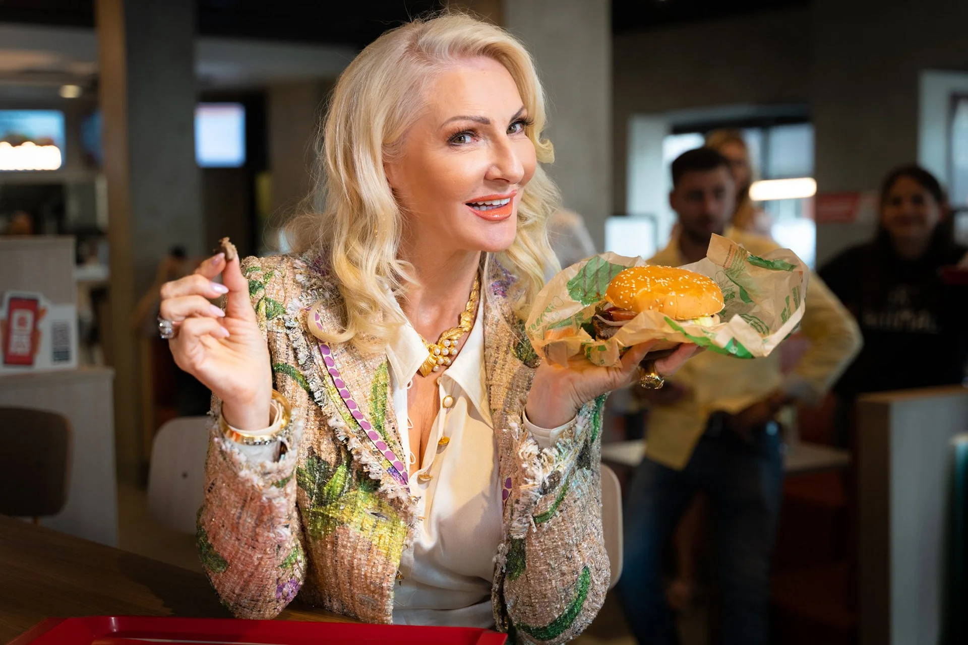 Eine blonde Frau in einem bunten Jackett hält einen Hamburger in der Hand und sitzt in einem Fast-Food-Restaurant, umgeben von mehreren Menschen im Hintergrund.