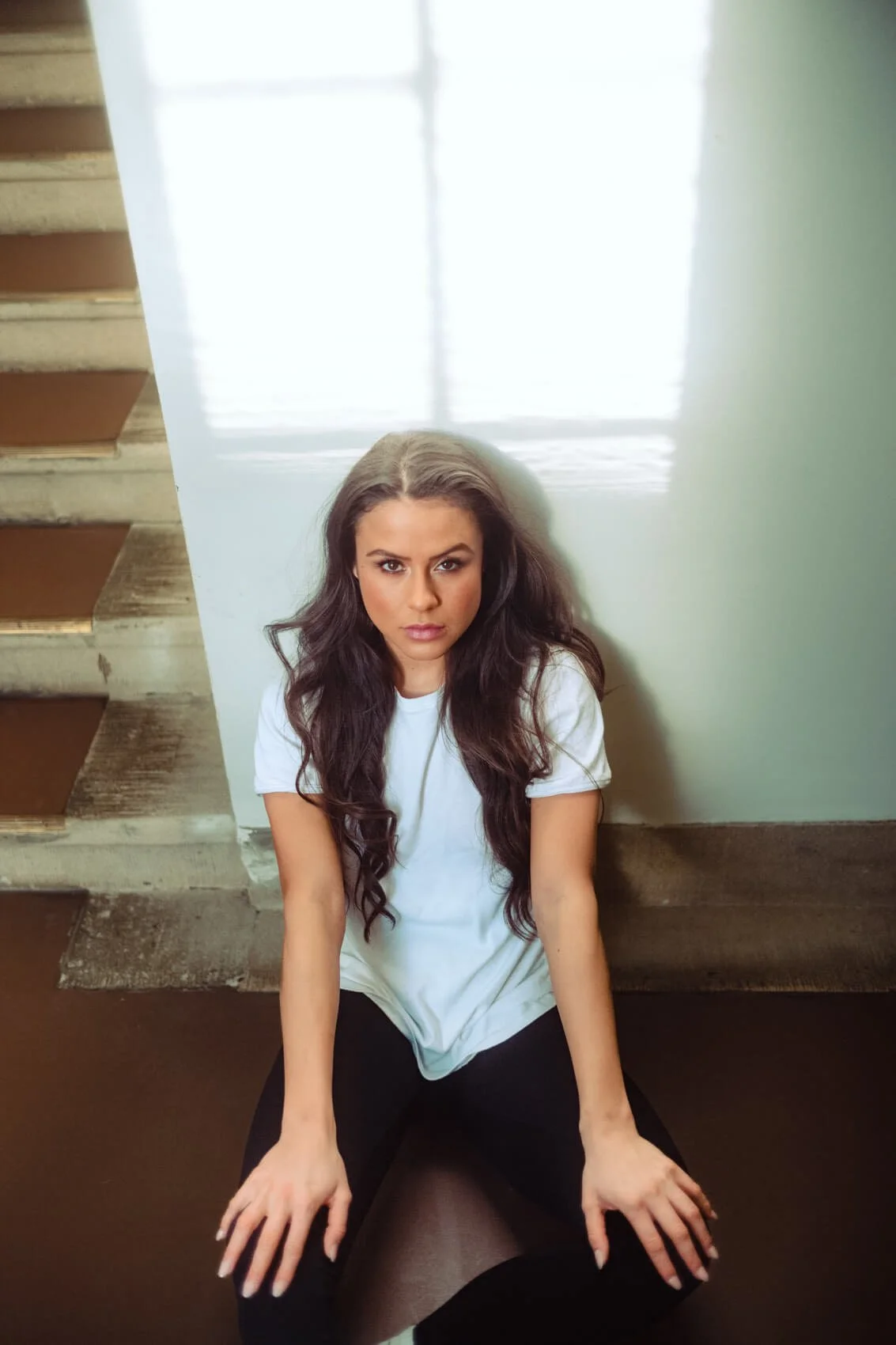 Frau mit langen braunen Haaren sitzt auf dem Boden vor einer weißen Wand mit einem Fenster und Schatten. Es gibt eine alte Holztreppe links.