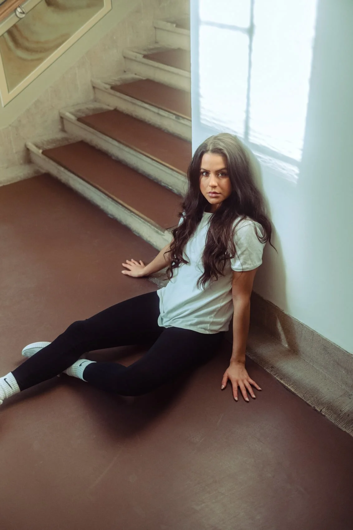Junge Frau sitzt an der Ecke einer Treppe auf dem Boden, lehnt an der Wand, mit dunklen, langen Haaren, weißem T-Shirt, schwarzen Hosen, weißen Socken, heller Wand und Treppe im Hintergrund.