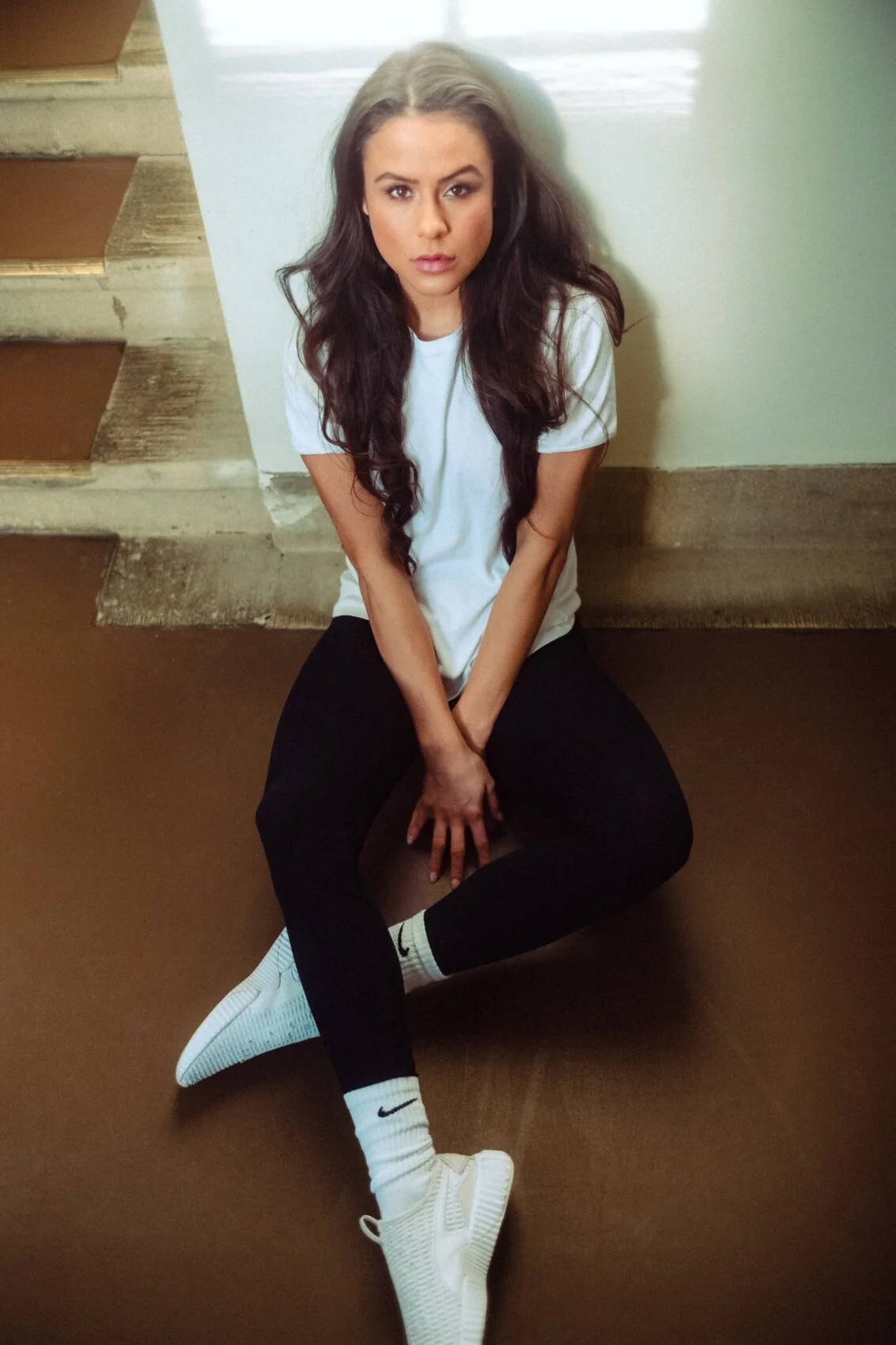 Eine junge Frau mit langen, braunen, lockigen Haaren sitzt auf dem Boden vor einer weißen Wand, trägt ein weißes T-Shirt, schwarze Hosen, weiße Nike-Socken und weiße Sportschuhe.