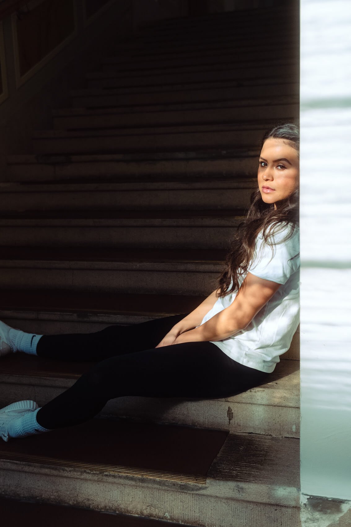 Junge Frau mit langen dunklen Haaren, sitzend an einer Wand neben Treppen, trägt weiße Sportschuhe, schwarze Leggings und ein weißes T-Shirt, schaut in die Kamera.