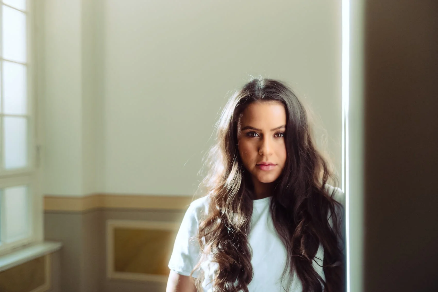 Junge Frau mit langen dunklen Haaren, die an einer Wand steht und in die Kamera schaut, in einem hell erleuchteten Raum.