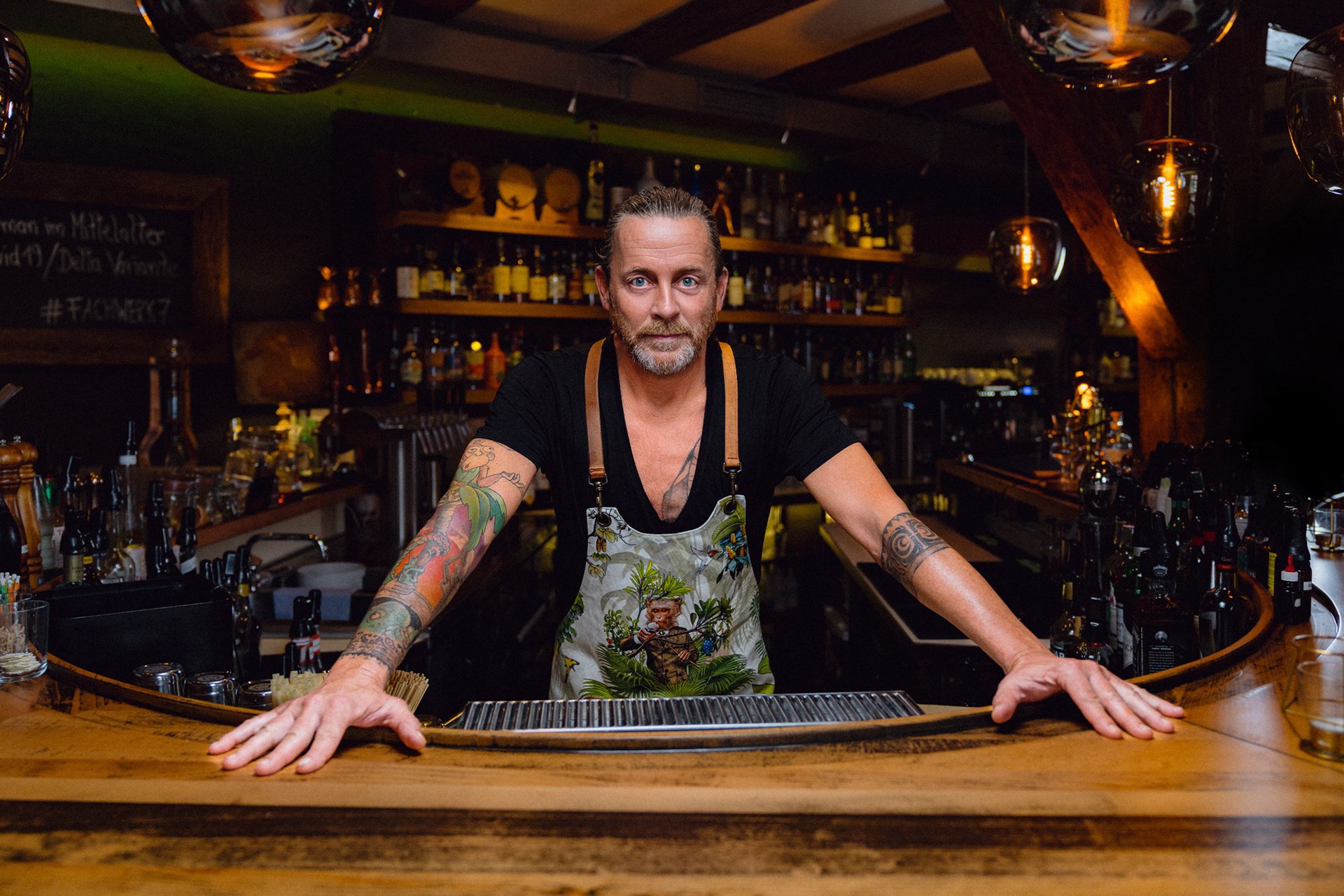 Ein bärtiger Mann mit Tattoos an den Armen steht hinter einer Bar, trägt ein schwarzes T-Shirt und eine farbenfrohe Schürze, im Hintergrund sind Regale mit Flaschen.