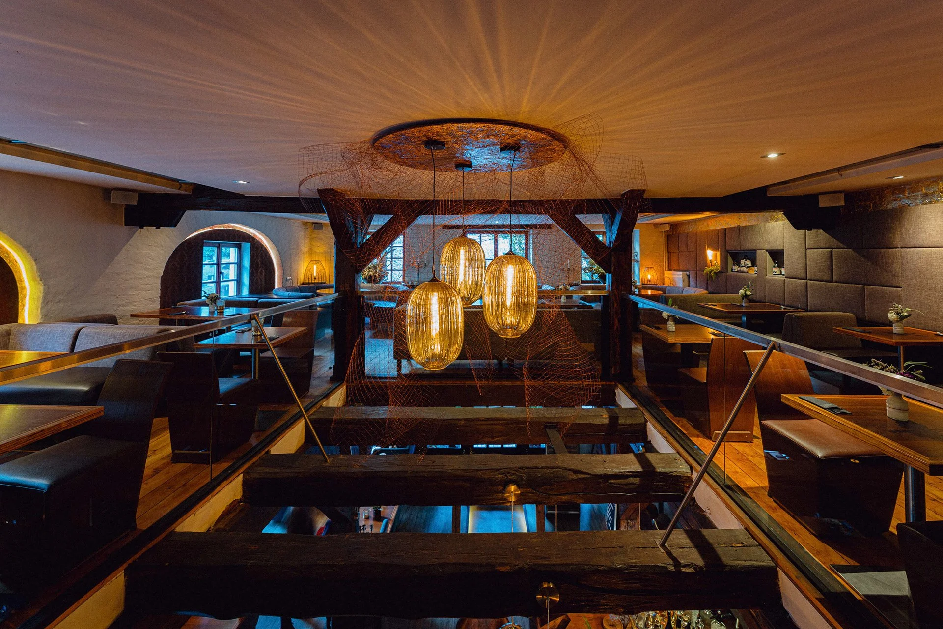 Innenansicht eines stilvollen Restaurants mit dunklen Holztischen, weichen Sitzbänken und warmem Licht, beleuchtet durch drei große, ovale goldene Lampen, mit Fenstern an den Wänden und dekorativen Elementen.