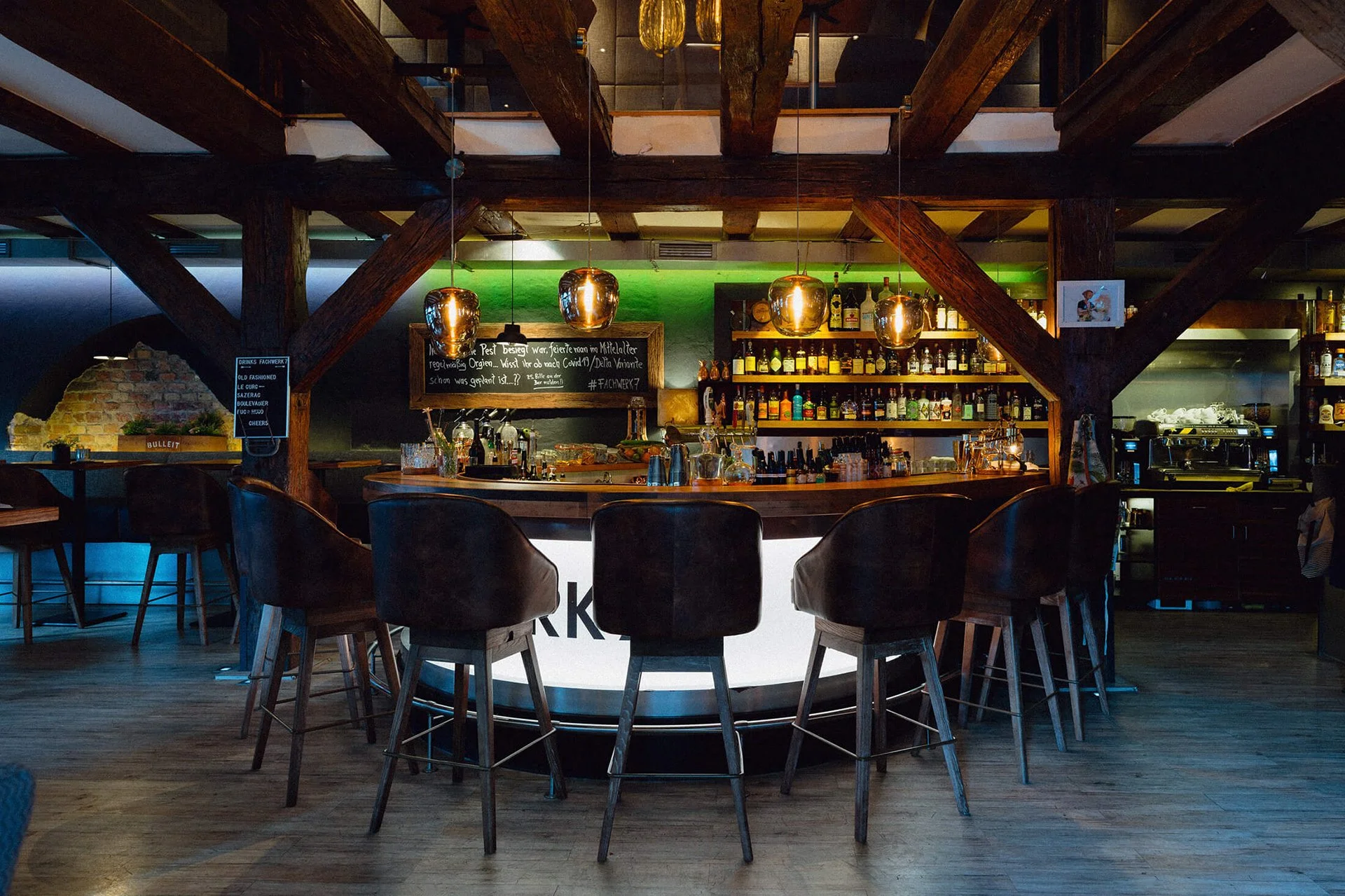 Innenaufnahme einer Bar mit dunkler Holztheke, vier Hockern, und einer Wand voller Spirituosen. Moderne Pendelleuchten hängen über der Bar, im Hintergrund sind weitere Sitzbereiche sichtbar.